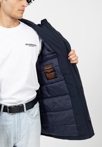 Marineblauer Puffermantel mit Reißverschluss, der ein gestepptes Innenfutter und ein orangefarbenes Etikett zeigt. Trägt man über einem weißen T-Shirt und Jeans.