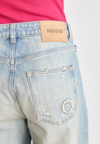 Artimame matomi šviesiai mėlyni nusidėvėję džinsai su kišenėje apvaliu plyšiu ir rusvai geltonu „HUGO“ ženkleliu viršuje, dėvimi su rausvu baltos spalvos viršumi.