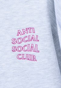 Jasnoszara bluza z różowym haftem, na którym widnieje napis „ANTI SOCIAL SOCIAL CLUB”. Tekst jest w zabawnym, nieregularnym kroju czcionki.
