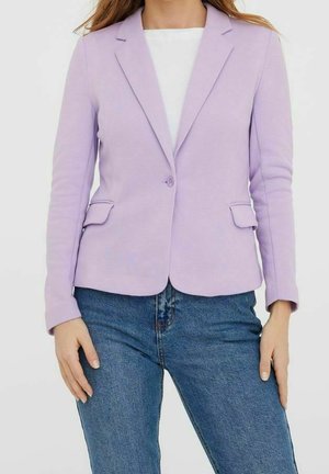 Kvinde iført en tætsiddende lilla blazer med en enkelt knap og klaplommer, kombineret med blå denimjeans og en hvid top under.