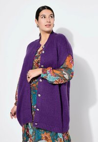 Lila gestrickte Strickjacke mit geripptem Saum, fünf silbernen Knöpfen und weiten Armausschnitten, die über einem bunten Kleid mit Blumenmuster getragen wird.