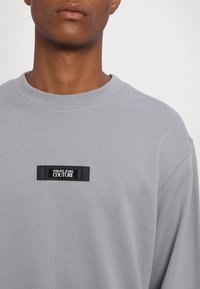 Grå sweatshirt med ribbat krage, prydd med en svart rektangulär logotyplapp som säger "VERSACE JEANS COUTURE." Slät textur, avslappnad design.