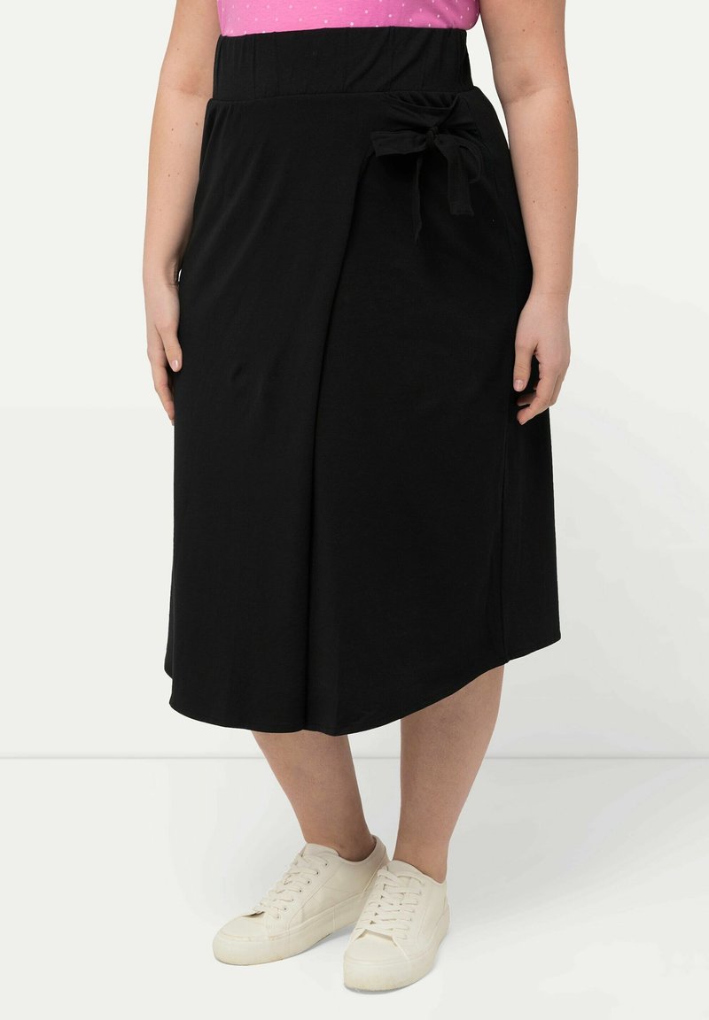 Zwarte wikkelrok met een strikdetail in de taille, gemaakt van zachte stof. Ggedragen met witte sneakers, met een vloeiende pasvorm en een lengte tot halverwege de kuit.