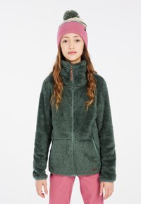Giacca di pelliccia sintetica verde con colletto alto e chiusura con zip, caratterizzata da una consistenza morbida e tasche frontali. Indossata con un cappello di lana rosa.