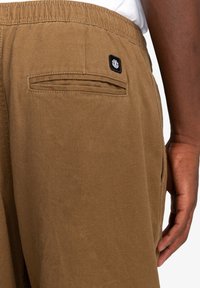 Bruine katoenen broek met een gestructureerde afwerking, voorzien van een achterzak en een klein zwart logo-opdruk. De tailleband is elastisch voor extra comfort.