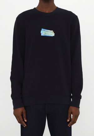 Person trägt ein schwarzes Langarm-Sweatshirt mit einem kleinen blauen und beigen Grafiklogo auf der Brust, auf dem "PS Paul Smith" steht.