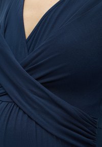 Κοντινό πλάνο ενός ατόμου που φοράει ένα σκούρο μπλε ένδυμα τύπου wrap με βαθύ V-λαιμόκοψη και ραμμένο ύφασμα στη μέση.