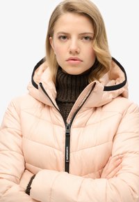 Giacca imbottita rosa con cappuccio bordato in nero, dotata di zip frontale e texture trapuntata. Indossata sopra un dolcevita marrone a coste.