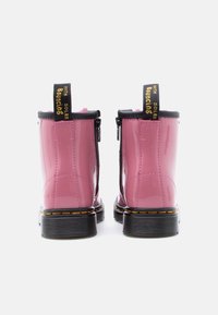 Γυαλιστερά ροζ μποτάκια αστραγάλου με μαύρες σόλες, κίτρινη ραφή και μαύρες θηλιές με την επιγραφή «ΜΕ ΕΛΑΣΤΙΚΕΣ ΣΟΛΕΣ», εμφανιζόμενα από πίσω σε λευκό φόντο.