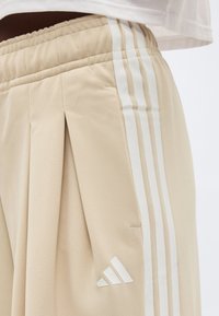 Μπεζ παντελόνι φόρμας Adidas με ελαστική μέση, λευκές τρεις ρίγες στο πλάι και λογότυπο Adidas κοντά στην τσέπη.