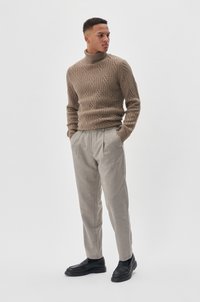 Pletivač turtleneck pulover u teksturiranoj bež boji, uparen s svijetlosivim nabranim hlačama i crnim mokasinama. Model stoji s rukama u džepovima.
