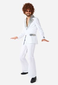 Costume de disco blanc avec des revers et des poches pailletés, pantalons évasés, porté avec des chaussures noires. Le mannequin a de grandes boucles de cheveux et des lunettes de soleil.
