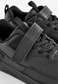 Baskets de sport noires présentant une combinaison de cuir lisse et texturé, avec une sangle Velcro, des lacets et des accents en tissu pour le soutien.
