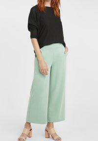 Svart, texturerad, löst sittande tröja i kombination med ljus mintgröna vida byxor. Modellen bär beige klacksandaler med blockklack.