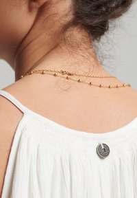 Top de tela blanca con tirantes finos, que presenta un botón plateado en el escote y un collar de cadena dorada con varias capas y pequeñas cuentas redondas.