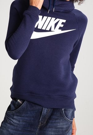 Una persona che indossa un hoodie blu navy con un grande logo bianco della Nike, abbinato a jeans in denim, che si copre parzialmente il viso con una mano.
