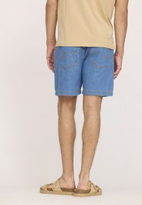 Shorts en denim bleu clair avec une coupe droite, présentant deux poches arrière et des détails surpiqués. Portés avec des chaussures beiges et une chemise beige.