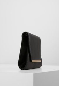Sac à main en cuir noir avec un rabat pliant, orné d'un accent en métal doré à l'avant, et une texture lisse.