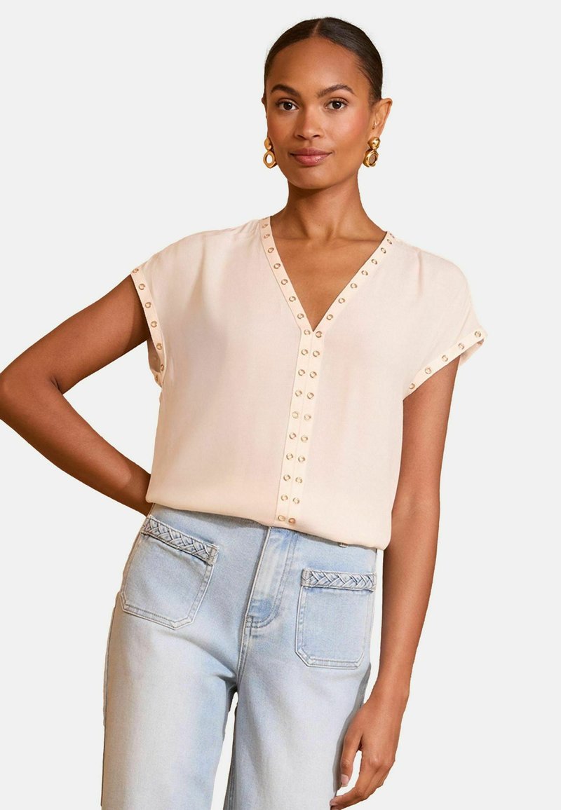 Blusa rosa pallido con scollatura a V, maniche corte e borchie circolari decorative lungo la pattina. Indossata con jeans blu chiaro a vita alta.