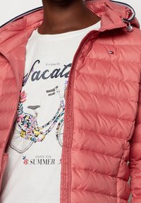 Doudoune rose clair avec fermeture éclair frontale et capuche, présentant une texture matelassée. En dessous, un t-shirt blanc avec un motif de vélo floral.