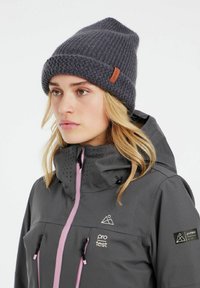 Graue Strickmütze mit umgeschlagenem Rand, die ein strukturiertes Muster aufweist und ein kleines braunes Leder-Brandlabel hat. Getragen mit einer grauen Jacke.