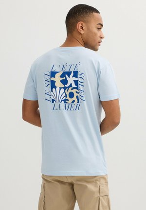 Hombre con camiseta azul claro con un gráfico de temática marina en azul y beige y texto en francés en la parte trasera, lleva pantalones cargo beige, de perfil.