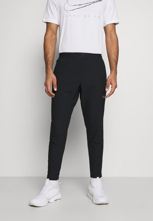 Mann trägt schwarze Nike Sporthosen, weiße Nike Sneakers und ein weißes Nike T-Shirt und steht vor einem schlichten Hintergrund.