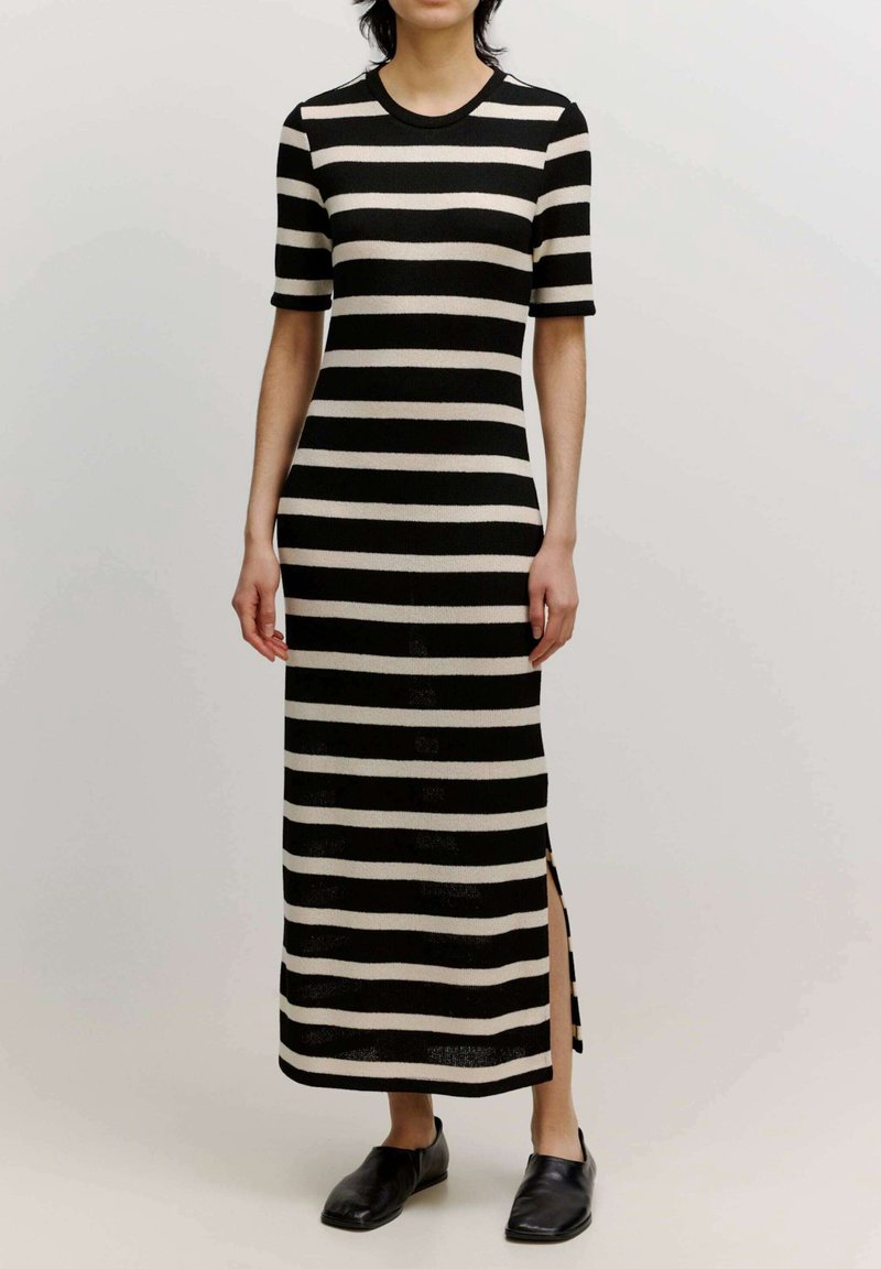 Personne portant une robe midi à rayures horizontales noires et beiges avec des manches courtes et des chaussures noires sans lacets, debout devant un fond uni.