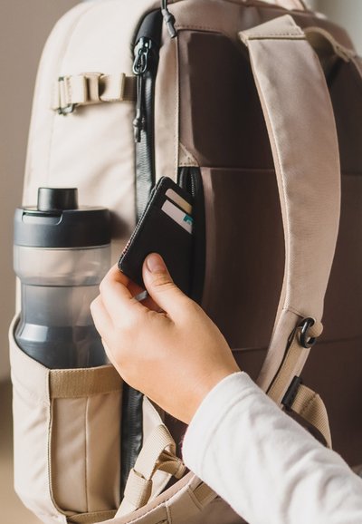 Mochila de tela beige con un bolsillo lateral para una botella de agua. Una mano saca una cartera negra con tarjetas de un compartimento con cremallera.
