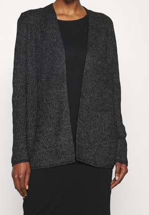 Personne portant un cardigan en maille noire sur une robe noire, debout devant un fond blanc uni, les mains détendues sur les côtés.