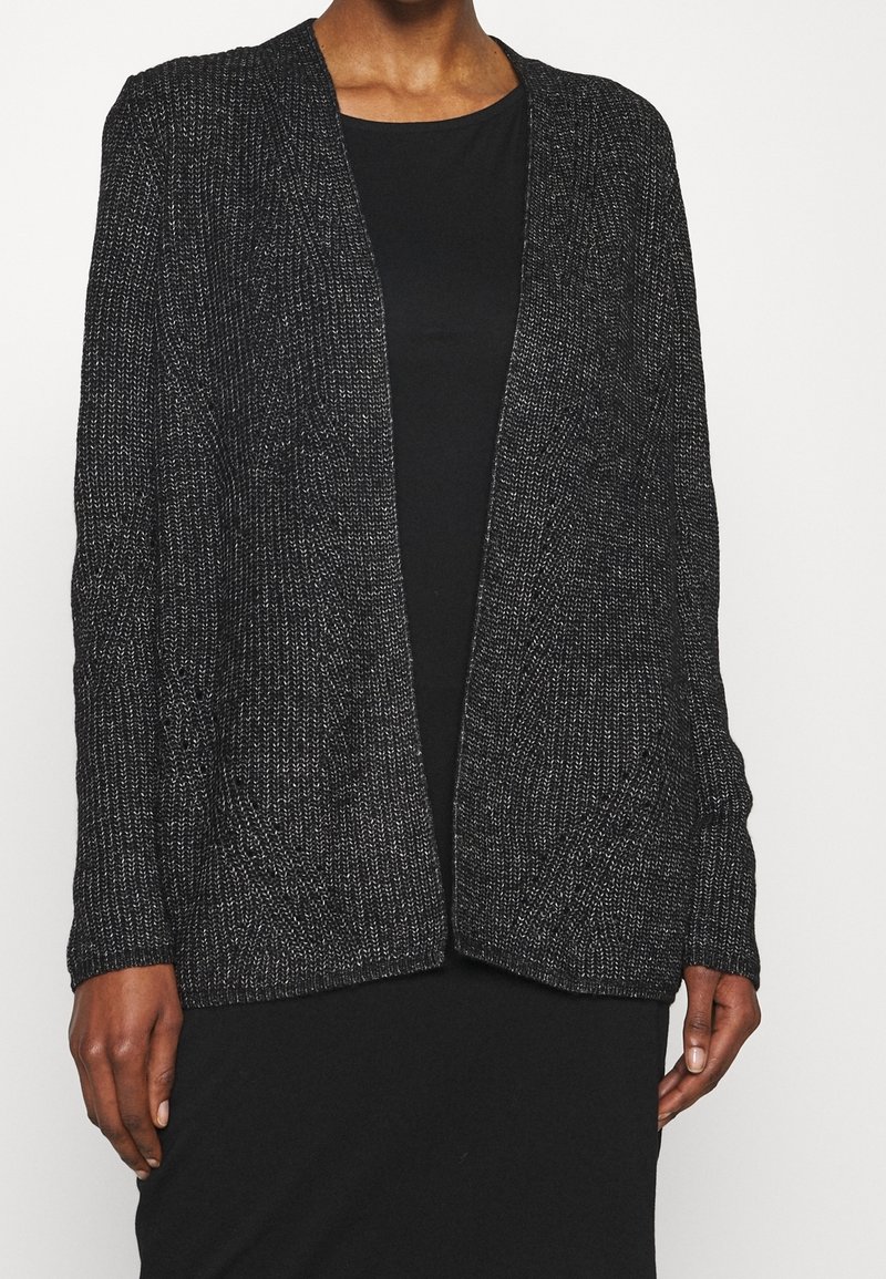 Personne portant un cardigan en maille noire sur une robe noire, debout devant un fond blanc uni, les mains détendues sur les côtés.