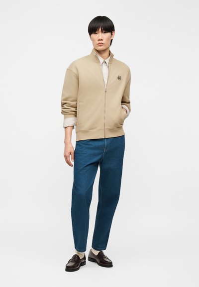 Νεαρός άνδρας με ίσια μαύρα μαλλιά στέκεται φορώντας μπεζ μπουφάν με φερμουάρ, μανίκια γυρισμένα, μπλε τζιν, άσπρες κάλτσες και σκούρα καφέ μοκασίνια.