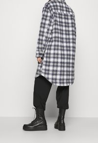 Camicia a quadri in navy e bianco, a maniche lunghe con orlo arrotondato. Pantaloni neri a sigaretta e stivaletti neri con lacci e suola spessa indossati.