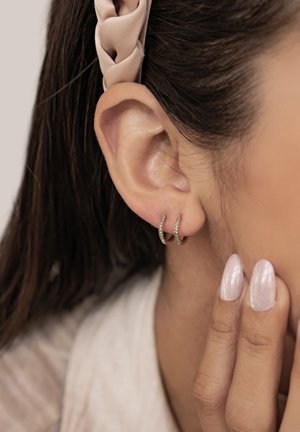 Roségoldene Creolen mit kleinen grünen Kristallen. Das Bild zeigt eine Nahaufnahme der Ohrringe, die an einem Ohr über glatter Haut getragen werden.