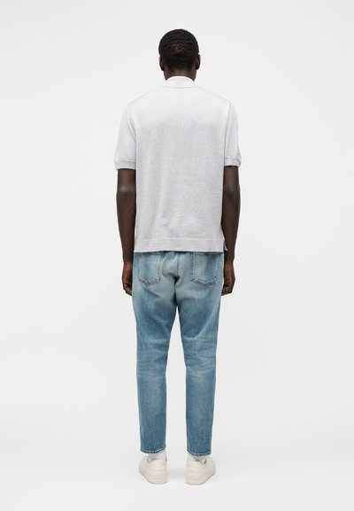 Άνδρας με πλάτη, που φορά ανοιχτογκρι κοντομάνικο πουκάμισο, μπλε τζιν, λευκές κάλτσες και λευκά αθλητικά παπούτσια μπροστά σε απλό φόντο.