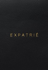 Superficie de cuero negro con el texto dorado en relieve "EXPATRIÉ." La textura es suave con un sutil patrón de grano.
