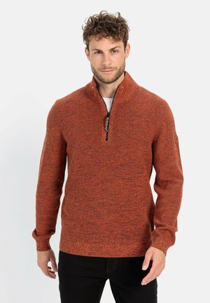 Strickpullover in strukturiertem verbranntem Orange mit einem halben Reißverschluss-Kragen, gerippten Bündchen und einem Patch auf dem linken Ärmel. Über einem weißen T-Shirt getragen.