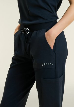 Persona che indossa pantaloni neri con coulisse in vita e logo visibile "FREDDY", mani nelle tasche anteriori, su uno sfondo semplice.