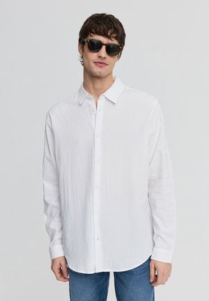 Giovane uomo con occhiali da sole neri, camicia bianca allentata con bottoni e jeans blu su uno sfondo grigio chiaro uniforme.