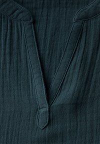 Tessuto verde scuro con una texture a coste, caratterizzato da una scollatura a V e un piccolo intaglio triangolare. Le cuciture lungo i bordi sono visibili.