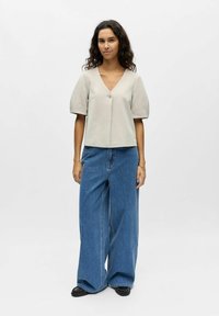 Blouse courte beige avec manches courtes bouffantes et col en V, dotée d'un bouton central. Associée à un pantalon large en denim bleu lâche.