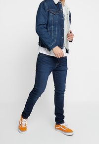 Jeansjacka med sherpalinning på kragen, mörkblå jeans och orange sneakers med vita detaljer, alla avbildade i stående pose.