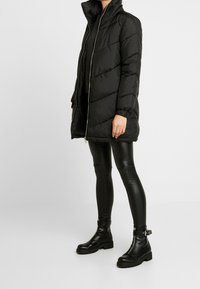 Abrigo de plumas negro con un cuello alto y costuras horizontales, combinado con leggings ajustados de piel sintética negra y botines.
