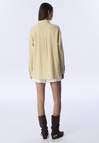 Camisa amarilla texturizada de corte holgado, con mangas largas y cuello abotonado, combinada con pantalones cortos blancos y botas marrones de caña baja.