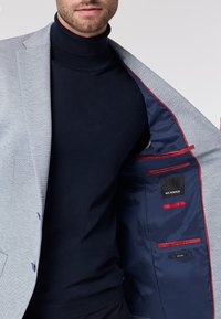 Blazer grigio chiaro con un motivo testurizzato, fodera blu e dettagli rossi. Collo alto blu navy sotto, che mette in risalto un design aderente.