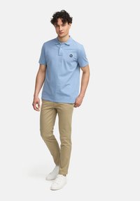 Lys blå polo skjorte med korte ermer og liten svart logo, sammen med beige bukser og hvite joggesko, stående på en hvit bakgrunn.