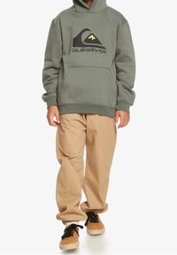 Green hoodie with Quiksilver logo, kangaroo pocket, ribbed cuffs, paired with beige pants and brown sneakers. Soft fabric, relaxed fit.