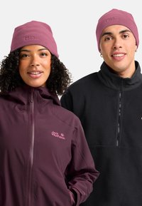 Twee mensen in Jack Wolfskin fleece jassen en bijpassende rozenkleurige mutsen, glimlachend voor een effen achtergrond.