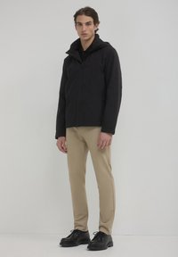 Giacca nera con zip e cappuccio, abbinata a pantaloni beige slim-fit e scarpe nere con lacci. Design semplice, texture del tessuto liscia.