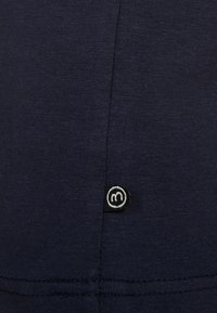 Primer plano de tela azul oscura con una costura vertical y una pequeña etiqueta cuadrada negra con un símbolo circular blanco.